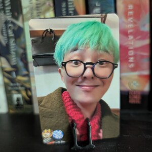 May include: A photo card featuring a person with green hair, wearing glasses and a red scarf. The card is propped up by a black stand. Books are visible in the background.