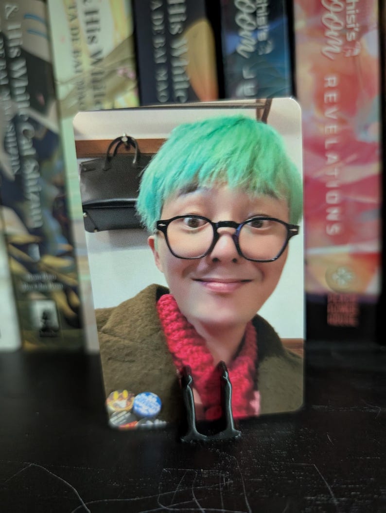 May include: A photo card featuring a person with green hair, wearing glasses and a red scarf. The card is propped up by a black stand. Books are visible in the background.