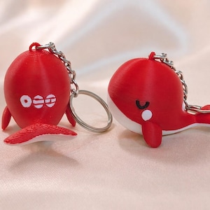 May include: Two red whale-shaped keyrings. One whale has the letters "OEE" in white, the other has a white belly and a small white dot. Both have silver keyrings and chains.