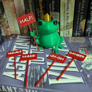 May include: A green frog figurine wearing a gold crown, holding a sign that says "HALP!". Several red signs with phrases like "TROUBLE" and "DANGER" are nearby. The frog is on a book shelf.