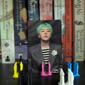 May include: A photo card featuring a person with green hair, wearing a gray blazer over a striped shirt, and a white flower pin. The card is propped up by a pink stand, with other colored stands in the foreground. Bookshelves are in the background.