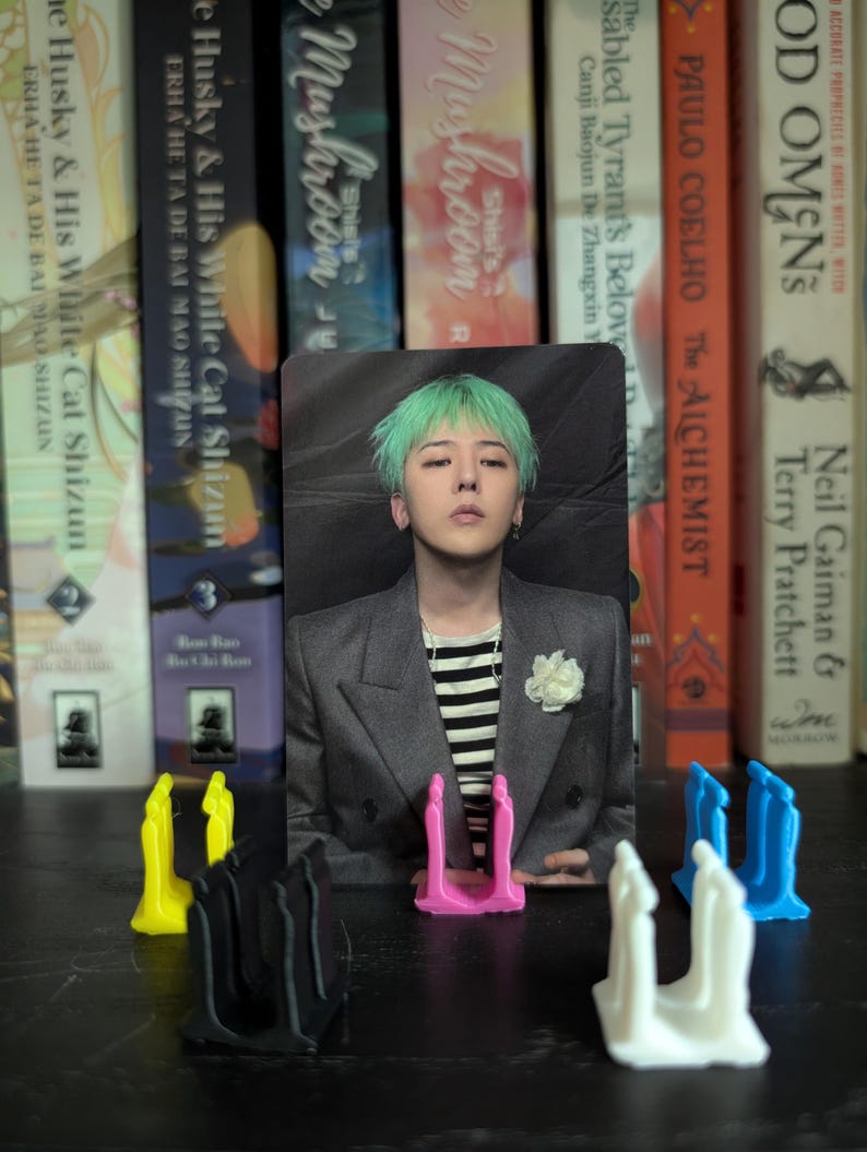 May include: A photo card featuring a person with green hair, wearing a gray blazer over a striped shirt, and a white flower pin. The card is propped up by a pink stand, with other colored stands in the foreground. Bookshelves are in the background.