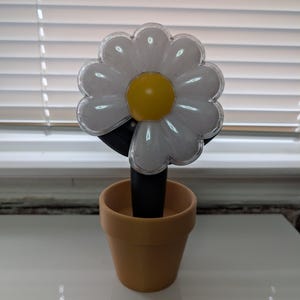 May include: A daisy-shaped lamp with a white, petal-like design and a yellow center. The lamp is mounted on a black stem and placed in a small, terracotta-colored flower pot. The lamp is designed to look like a flower.