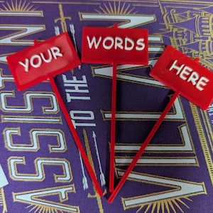 Puede incluir: Tres letreros rojos con texto blanco "YOUR", "WORDS" y "HERE". Los letreros están sobre finos tallos rojos. El fondo es una portada de libro morada con letras doradas y una pequeña etiqueta blanca.