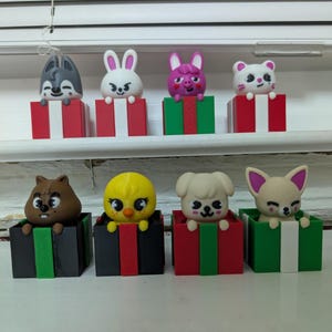 Könnte beinhalten: Eine Sammlung von acht farbenfrohen Tierfiguren, die jeweils aus einer dekorativen Geschenkbox auftauchen. Die Figuren umfassen einen Wolf, ein Kaninchen, ein Schwein, einen Bären, einen Biber, ein Küken, einen Hund und einen Fuchs. Die Boxen sind rot, grün und schwarz.