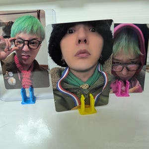 May include: Three photo cards featuring a person with green hair and glasses. The cards are held in clear plastic holders and supported by blue, yellow, and pink card stands. The person is wearing different outfits in each photo.