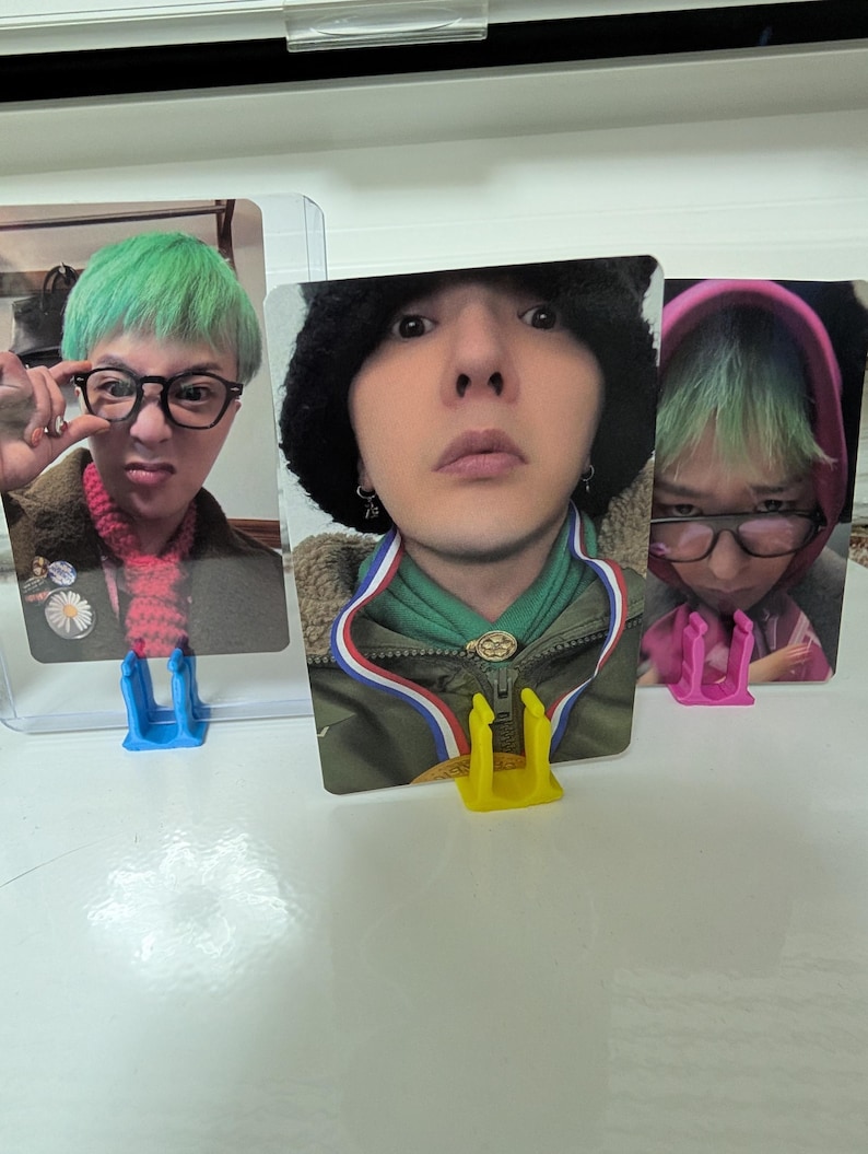 May include: Three photo cards featuring a person with green hair and glasses. The cards are held in clear plastic holders and supported by blue, yellow, and pink card stands. The person is wearing different outfits in each photo.