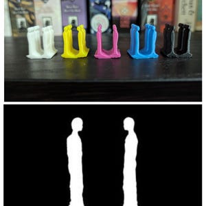 May include: A row of colorful bookends in white, yellow, pink, blue, and black. Each bookend has a unique design, with a U-shaped structure and a silhouette of two figures. The bookends are on a dark surface with books in the background.