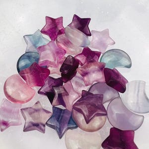 May include: A collection of star and moon-shaped gemstones in shades of purple, pink, and blue. The polished stones vary in size and are arranged on a white surface. The gemstones are likely used for jewelry or decorative purposes.