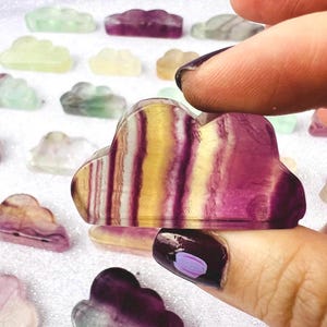 May include: A collection of cloud-shaped gemstone carvings in various colors, including purple, yellow, and green. The focus is on a cloud-shaped carving held between fingers, showcasing a striped pattern of purple and yellow. The background features more cloud carvings.