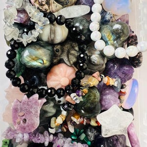 May include: A collection of various gemstones and beaded bracelets in a clear container. The gemstones are in shades of purple, green, black, and white. Some stones are carved into shapes like stars and flowers. The bracelets feature black and white beads.