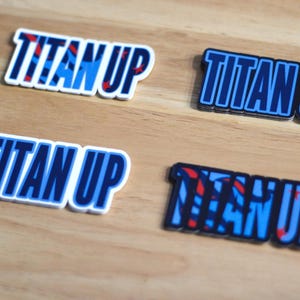 May include: Four stickers with the text "TITAN UP" in blue and white lettering on a white background. The stickers have different designs and patterns.