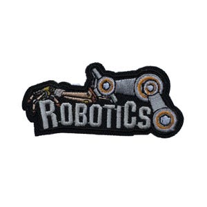 May include: Embroidered patch with the word "ROBOTICS" in white block letters. The patch features a stylized robot design in silver, gold, and brown, set against a black background. The patch is likely for clothing or accessories.