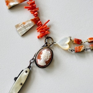 Può includere: Una collana con un ciondolo cammeo ovale centrale con un profilo bianco su sfondo marrone, accentuato da un ciondolo in madreperla a forma di cuore, un lungo pezzo di conchiglia bianca e fili di rami di corallo arancione brillante e perline rettangolari.