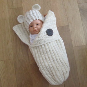 Puede incluir: Un capullo de bebé de ganchillo color crema con un gorro a juego con orejas de oso. El capullo se cierra con un botón grande y oscuro. El bebé está envuelto en el capullo, solo se ve la cara.