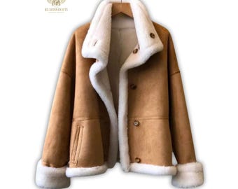 May include: A tan suede jacket featuring a thick white faux shearling lining, a wide notched collar, and long sleeves with white shearling cuffs. The jacket includes a front button closure with brown buttons and two vertical slit pockets.