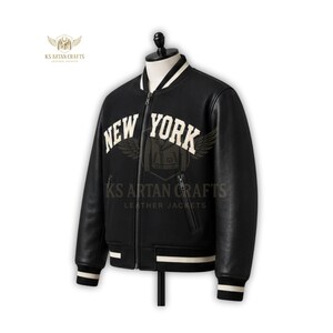 May include: A black leather bomber jacket with the text "NEW YORK" in white block letters across the chest. The jacket has white stripes on the collar, cuffs, and hem. The jacket is displayed on a mannequin.