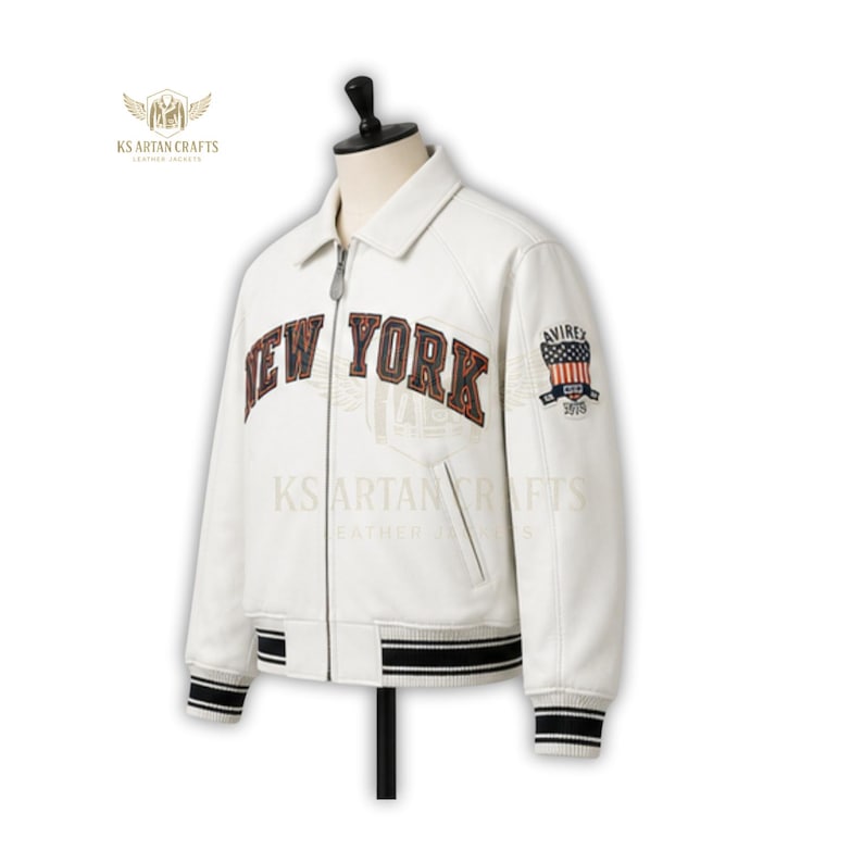 May include: A white leather jacket with the words "NEW YORK" across the chest in a brown, distressed font. The jacket has a collar, a zipper closure, and black and white striped cuffs and hem. An American flag patch is on the sleeve.