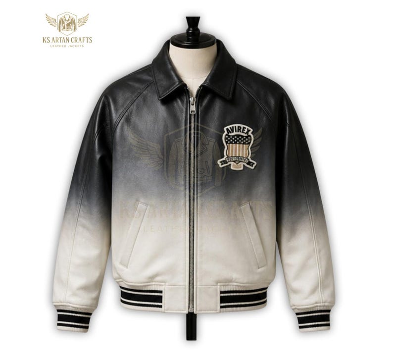 K&ouml;nnte beinhalten: Eine schwarz-wei&szlig;-Ombre-Lederjacke mit Rei&szlig;verschluss vorne und geripptem Kragen und Manschetten. Die Jacke hat einen Aufn&auml;her auf der Brust mit dem Text "AVIREX" und einem amerikanischen Flaggen-Design. Die Jacke wird auf einer Schaufensterpuppe pr&auml;sentiert.