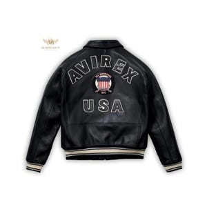 Könnte beinhalten: Schwarze Lederjacke mit den gestickten Schriftzügen "AVIREX" und "USA" auf dem Rücken. Die Jacke hat weiß-cremefarbene Streifen an den Manschetten und am Saum. Ein Aufnäher mit "ALL AMERICANS" und einer US-Flagge ist über "USA" zentriert.