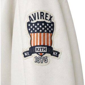 May include: Close-up of a white leather jacket featuring a patch with the text "AVIREX" above a design of stars and stripes. Below the design is the text "KITH" and "1975". The jacket has a classic design.