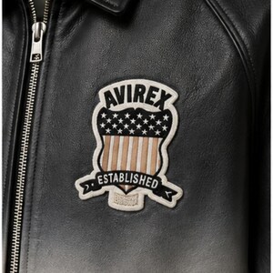 K&ouml;nnte beinhalten: Nahaufnahme einer schwarzen Lederjacke mit silbernem Rei&szlig;verschluss und einem Aufn&auml;her. Der Aufn&auml;her zeigt das Wort "AVIREX" &uuml;ber einem Schilddesign mit Sternen und Streifen und das Wort "ESTABLISHED" auf einem Banner.