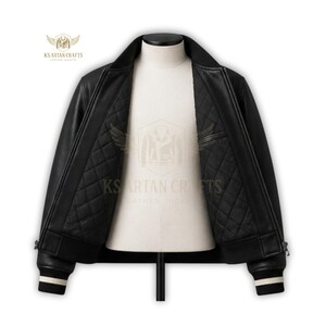 May include: Black leather bomber jacket with a quilted interior, displayed on a mannequin. The jacket features a ribbed collar, cuffs with white stripes, and a zipper closure. The interior lining is black and diamond-stitched.