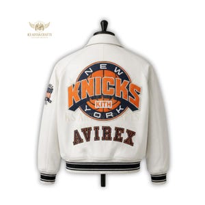 May include: Cream-colored varsity jacket with the New York Knicks logo in orange and navy blue on the back. The jacket features the Avirex brand name and a striped black and white ribbed hem. A small American flag patch is on the sleeve.