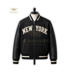 May include: Black leather varsity jacket with the text "NEW YORK" in white block letters. The jacket has a white collar, cuffs, and waistband with black stripes. The jacket is zipped up and has two front pockets.