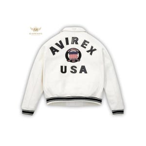 May include: White jacket with the word "AVIREX" arched across the back in black lettering, above "USA". A circular emblem with the American flag is centered between the words. The jacket has black and white striped trim on the cuffs and hem.