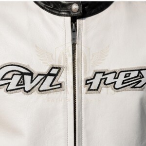 May include: Close-up of a white leather jacket with black accents on the collar and shoulders. The jacket features the embroidered text "Avi Nex" in black and white, with a gray outline. A silver zipper runs down the center.