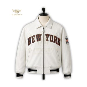 May include: White leather jacket with "NEW YORK" lettering across the chest. The jacket has a black and white striped hem and cuffs, a zipper closure, and a small patch with an American flag design on the sleeve.