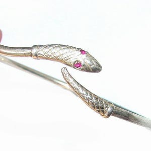 May include: A silver-toned open cuff bracelet shaped like a snake, featuring a textured scale pattern along the body and head. The snake head is detailed with two small, vibrant red circular stones set as eyes, creating a distinct focal point.