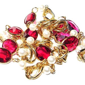 May include: A gold necklace with large, oval, ruby-red gemstones and creamy white pearls. The necklace has a chain of interlocking gold links. The jewellery is set against a white background.