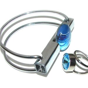 May include: A silver-toned bracelet and ring set. The bracelet features a blue, oval-shaped stone and a rectangular clasp. The ring has a similar blue stone and a double-band design. The jewelry is set against a white background.