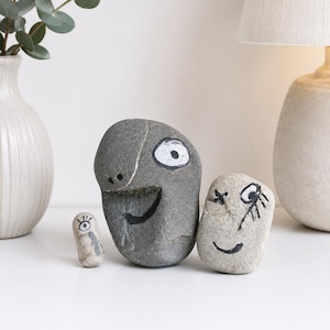 Puede incluir: Tres piedras de río lisas y redondeadas pintadas con rasgos faciales sencillos en blanco y negro. La piedra gris oscuro más grande tiene una sonrisa amplia y un ojo, la mediana gris claro tiene un ojo guiñando y la más pequeña tiene un solo ojo.