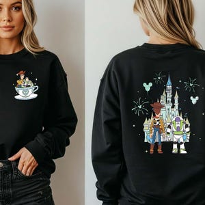 K&ouml;nnte beinhalten: Schwarzes Sweatshirt mit einem Cartoon-Design von Toy Story-Charakteren in einer Teetasse auf der Vorderseite und einem Schloss mit Charakteren auf der R&uuml;ckseite. Das Sweatshirt hat lange &Auml;rmel und einen Rundhalsausschnitt.
