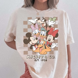 May include: Ivory t-shirt with a graphic of Mickey Mouse, Minnie Mouse, Donald Duck, Daisy Duck, Goofy, and Pluto. The design is set against a brown checkered background, with "Mickey & Co. Est. 1928" printed below. The shirt is paired with red and white checkered shorts.