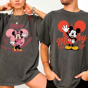Może przedstawiać: Dwie grafitowe koszulki z postaciami Disneya. Jedna koszulka przedstawia Minnie Mouse w r&oacute;żowej sukience w grochy z napisem "Minnie". Druga koszulka przedstawia Myszka Miki z napisem "Mickey 1928".