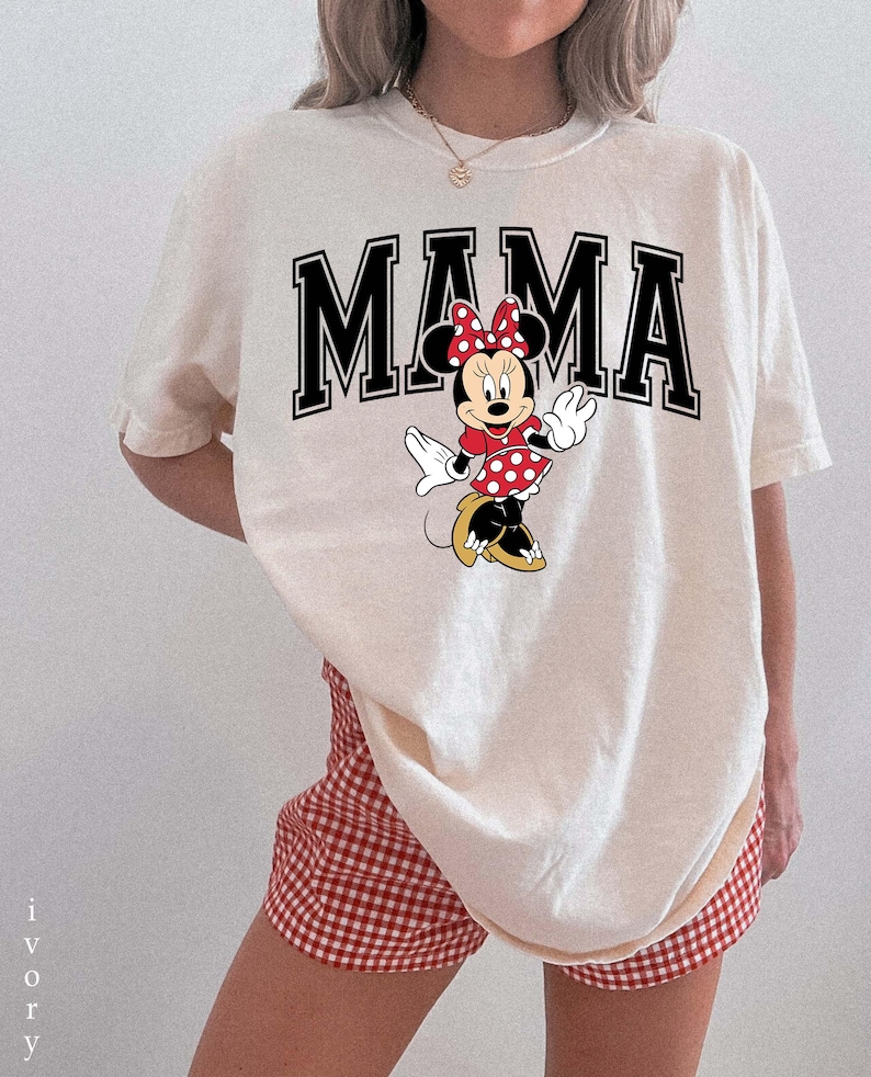 May include: Ivory-colored t-shirt with the word "MAMA" in black block letters, featuring a Minnie Mouse graphic. Minnie Mouse is wearing a red polka dot dress and bow. The model is wearing red and white checkered shorts.