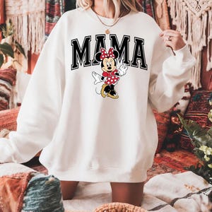 May include: White sweatshirt with the word "MAMA" in black block letters and a cartoon Minnie Mouse graphic. Minnie Mouse is wearing a red polka dot dress and bow. The sweatshirt is a casual style.