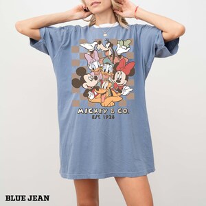May include: Blue jean-coloured t-shirt with a graphic of Mickey & Co. characters, including Mickey Mouse, Minnie Mouse, Donald Duck, and Goofy. The design is set against a brown and tan checkered background, with the text "Mickey & Co. Est. 1928".