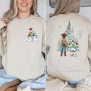 K&ouml;nnte beinhalten: Beiges Sweatshirt mit einem Cartoon-Design von Woody und Buzz Lightyear. Auf der Vorderseite sind sie in einer Teetasse zu sehen, w&auml;hrend die R&uuml;ckseite sie mit einem Schloss und Feuerwerk zeigt. Das Design ist in Blau-, Gr&uuml;n- und Braunt&ouml;nen gehalten.