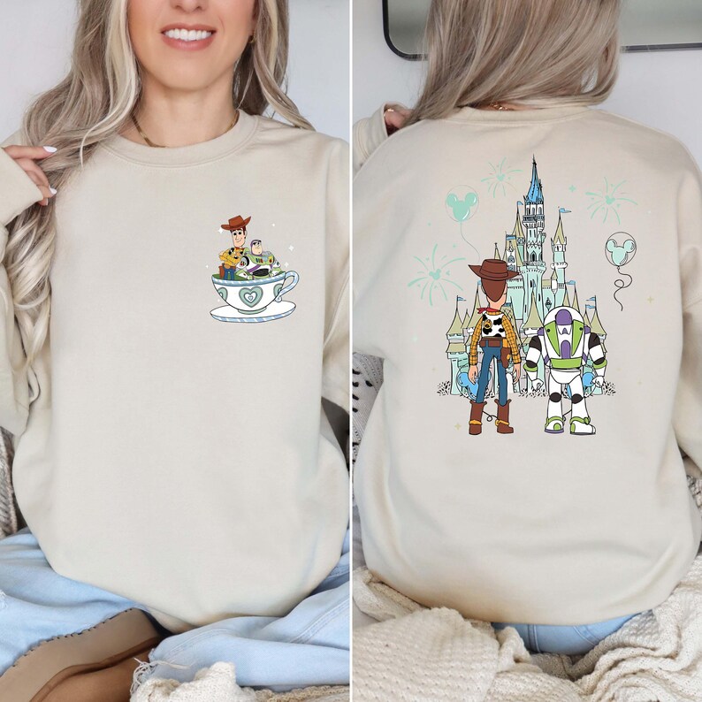 K&ouml;nnte beinhalten: Beiges Sweatshirt mit einem Cartoon-Design von Woody und Buzz Lightyear. Auf der Vorderseite sind sie in einer Teetasse zu sehen, w&auml;hrend die R&uuml;ckseite sie mit einem Schloss und Feuerwerk zeigt. Das Design ist in Blau-, Gr&uuml;n- und Braunt&ouml;nen gehalten.