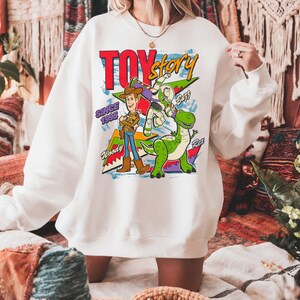 May include: White sweatshirt featuring a colorful graphic of Toy Story characters Woody, Buzz Lightyear, and Rex. The design includes the text "TOY STORY" in red and yellow, with "SINCE 1995" below. The sweatshirt has long sleeves.