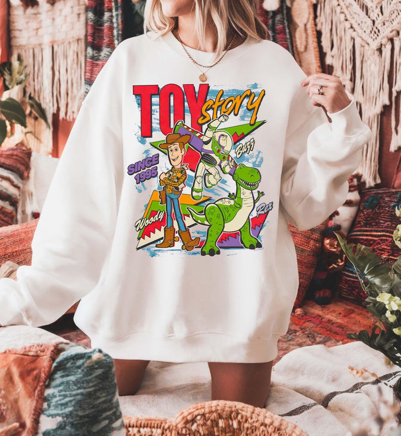 May include: White sweatshirt featuring a colorful graphic of Toy Story characters Woody, Buzz Lightyear, and Rex. The design includes the text "TOY STORY" in red and yellow, with "SINCE 1995" below. The sweatshirt has long sleeves.