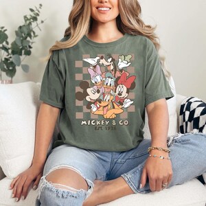 May include: Moss green t-shirt with a graphic of Mickey Mouse, Minnie Mouse, Donald Duck, Daisy Duck, Goofy, and Pluto. The shirt has the text "Mickey & Co. EST. 1928" in a retro font. The t-shirt is a relaxed fit.