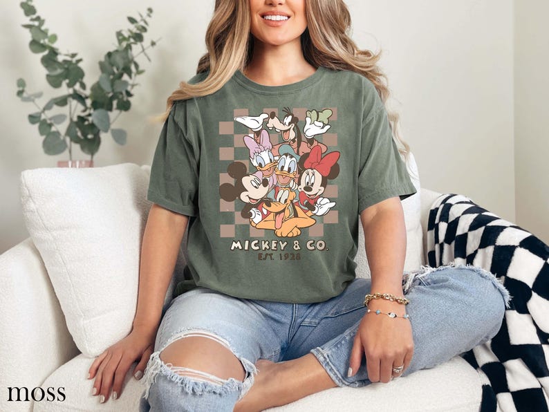 May include: Moss green t-shirt with a graphic of Mickey Mouse, Minnie Mouse, Donald Duck, Daisy Duck, Goofy, and Pluto. The shirt has the text "Mickey & Co. EST. 1928" in a retro font. The t-shirt is a relaxed fit.