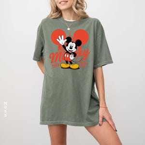 Może przedstawiać: Oliwkowo-zielona koszulka z grafiką Myszki Miki. Projekt zawiera Myszka Miki machającego, czerwone uszy i napis "Mickey" w czerwonym skrypcie. Rok "1928" jest r&oacute;wnież w kolorze czerwonym. Koszulka ma luźny kr&oacute;j.