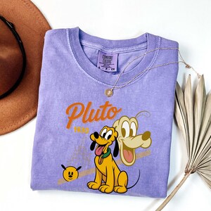 Może przedstawiać: Liliowa koszulka Comfort Colors z grafiką Plutona, psa z kresk&oacute;wek Disneya, oraz napisem "Pluto 1930". Projekt zawiera kresk&oacute;wkowy wizerunek Plutona i mały obraz twarzy Plutona. Złoty naszyjnik jest zarzucony na koszulkę.
